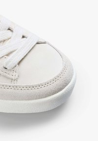 Beżowe zamszowe sneakersy z białymi sznurówkami i gumową podeszwą. Posiadają zaokrąglony czubek, detal w postaci przeszyć wzdłuż krawędzi oraz teksturowaną podeszwę.