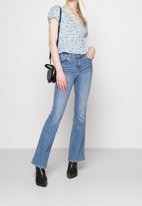 Blusa floreale blu con maniche a palloncino, abbinata a jeans a zampa d'elefante blu chiaro e stivali neri alla caviglia. Una borsa a tracolla nera aggiunge contrasto.