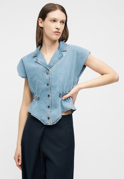 Femme portant un haut en denim bleu clair sans manches avec boutons et taille froncée, associée à un pantalon noir taille haute, debout avec une main sur la hanche.