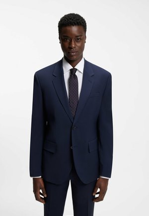Mann trägt einen dunkelblauen, taillierten Anzug mit weißem Hemd und schwarzer Krawatte mit kleinen weißen Punkten, stehend vor einem schlichten weißen Hintergrund.