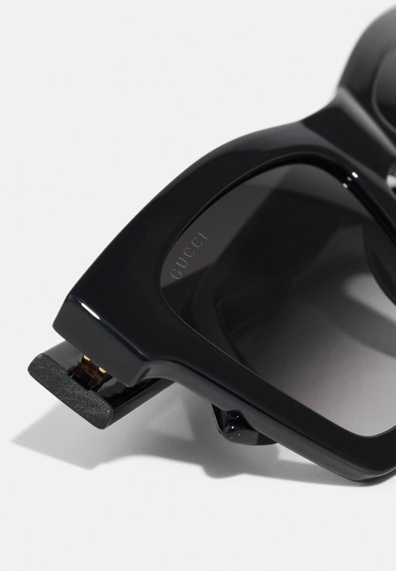 Lunettes de soleil noires Gucci avec une monture épaisse et angulaire, verres foncés dégradés, gravure subtile du logo et accents en métal doré.