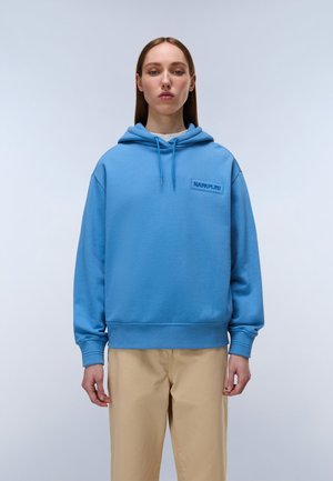 Junge Person mit glatten Haaren trägt einen blauen Kapuzenpullover mit "NAPAPIJRI"-Logo und beigen Hosen, steht vor einem einfarbigen hellen Hintergrund.
