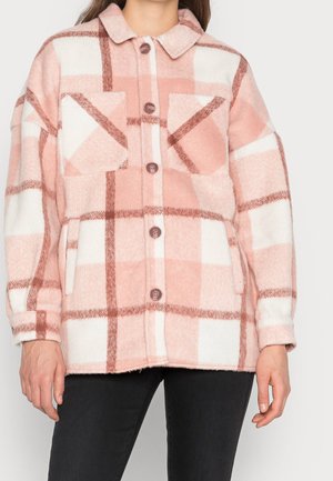 Kobieta w różowo-białej koszuli w kratę z kieszeniami na piersi i czarnych spodniach, stojąca na jednolitym tle.