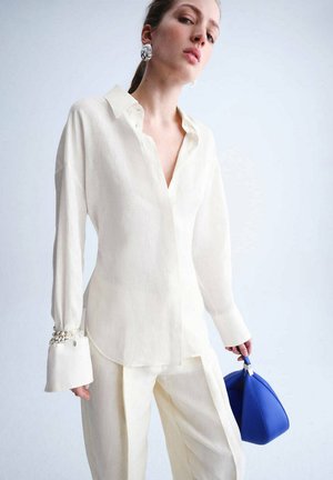 Frau in lockerem weißen Hemd und Hose, die eine blaue Handtasche hält, trägt große silberne Ohrringe und ein Kettenarmband am Ärmel.