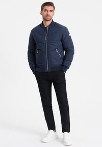 Námornícká zateplená bunda s vysokým límcem, předním zipem a dvěma bočními kapsami, v kombinaci s černými kalhotami a bílými teniskami.