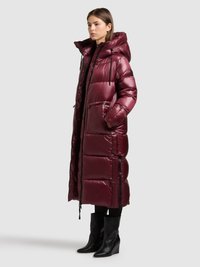 Lange bordeauxrode donsjas met capuchon, met een glanzende textuur, ritszakken en contrasterende zwarte ritsen, gecombineerd met zwarte sleehakken.