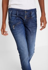 Siniset denimmit, joissa kapeahko leikkaus, vetoketjullinen tasku, useita nappisulkimia ja kontrastitikkaukset.