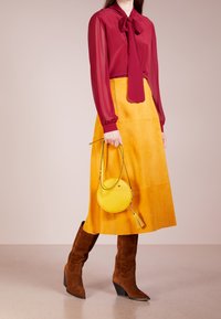 Donna che indossa una blusa traslucida bordeaux e una gonna midi gialla senape, mentre tiene una borsa rotonda gialla e indossa stivali alti fino al ginocchio in suede marrone.