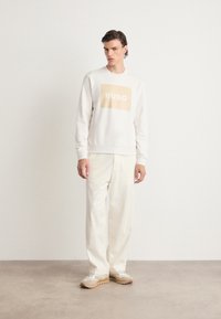 Weißer Hoodie mit "HUGO" in beigem Quadrat, kombiniert mit lockeren beigen Hosen und beigen Sneakers. Glatte Textur, lässige Passform, minimales Design.