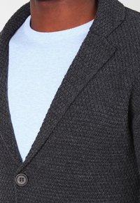 Cardigan lavorato a maglia grigio scuro con una finitura texturizzata, caratterizzato da un colletto a risvolto, due bottoni, indossato sopra una camicia azzurro chiaro.