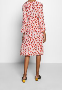 Frau in einem knielangen weißen Kleid mit roten Herzmustern, langen Ärmeln, einem Taillengürtel und schwarzen, niedrig-absatzschuhen mit gelben Absätzen.