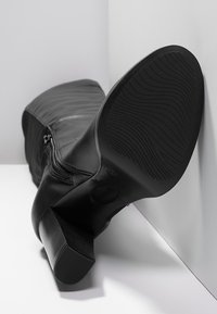 Stivale in pelle nera con tacco, con cerniera, posizionato in un angolo su una superficie bianca e grigia, con la suola rivolta verso la fotocamera.