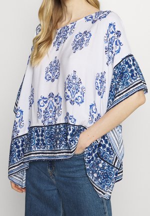 Mujer con una blusa holgada blanca y azul con estampado floral y mangas anchas y vaqueros azules, mano en el bolsillo.