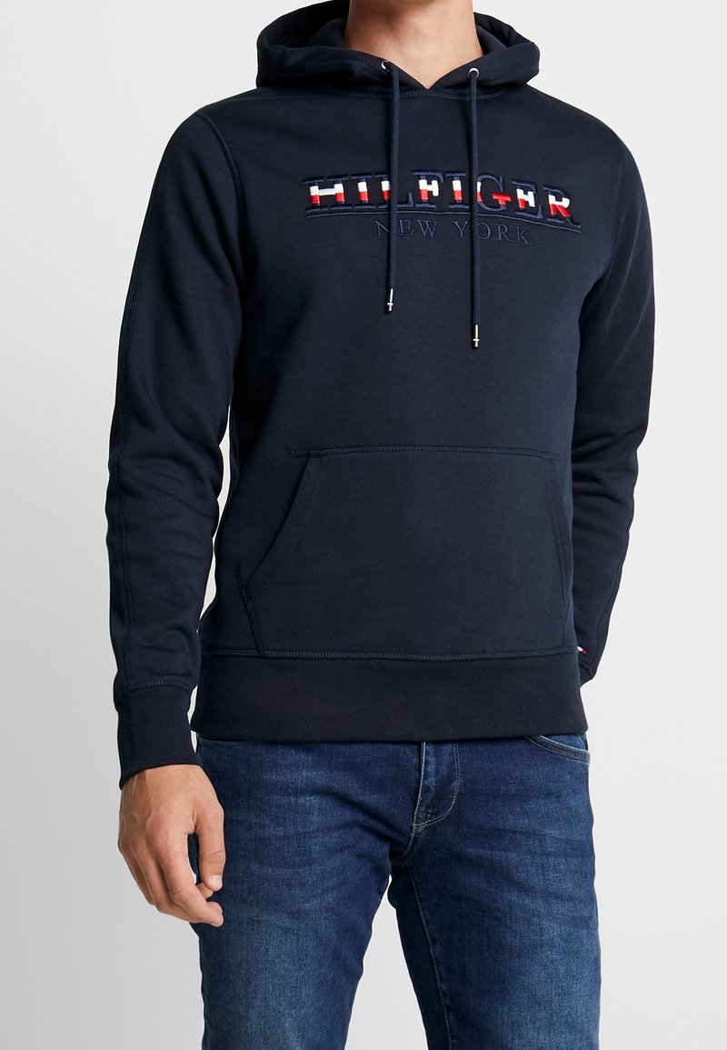 Sudadera marinera con bolsillo frontal tipo canguro, capucha con cordón y logo bordado en rojo, blanco y azul. Tela suave de mezcla de algodón.