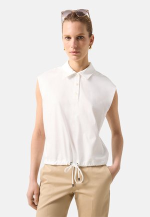 Kvinde iført hvid ærmeløs skjorte med krave og snøre ved taljen, beige bukser, gyldne øreringe og solbriller på hovedet.