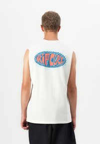 Rövid göndör hajú férfi, aki fehér ujjatlan pólót visel, hátán színes "Rip Curl" logóval, fekete sorttal párosítva.
