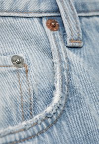 Lyseblåt denimstof med synlige syninger, to metalnitter og en flossede lommeoverkant.