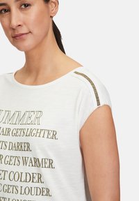 T-shirt en coton blanc à manches courtes, avec du texte et des accents métalliques dorés sur l'épaule, offrant une coupe décontractée et une texture douce.