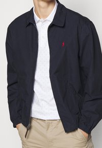 Marine lichte jas met een rits aan de voorkant, elastische manchetten en zijzakken, gedragen over een witte polo met blauwe stippen en een logo.