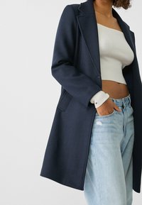 Marineblå frakke med skræddersyet design, markante revers og to forlommer. Båret over en hvid ribstrikket crop top og lyseblå slidte jeans.