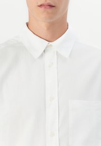 Nærbilde av en person som har på seg en hvit skjorte med krage og brystlomme mot en enkel hvit bakgrunn.