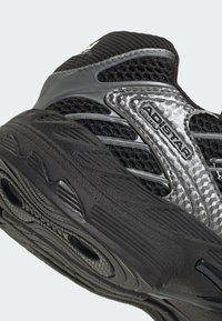 Čierna športová obuv z sieťovinového a syntetického materiálu s textúrovanou podrážkou, striebornými detailmi a výrazným logom na boku.