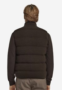 Dunkelbraune Pufferweste mit gerippter Struktur, hohem Kragen und dicken horizontalen Abschnitten. Über einem Langarmshirt getragen, das eine glatte Oberfläche zeigt.