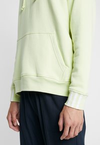 Světle zelená bavlněná mikina s klokaní kapsou, s bílými pruhovanými akcenty na manžetách a žebrovaném lemu.