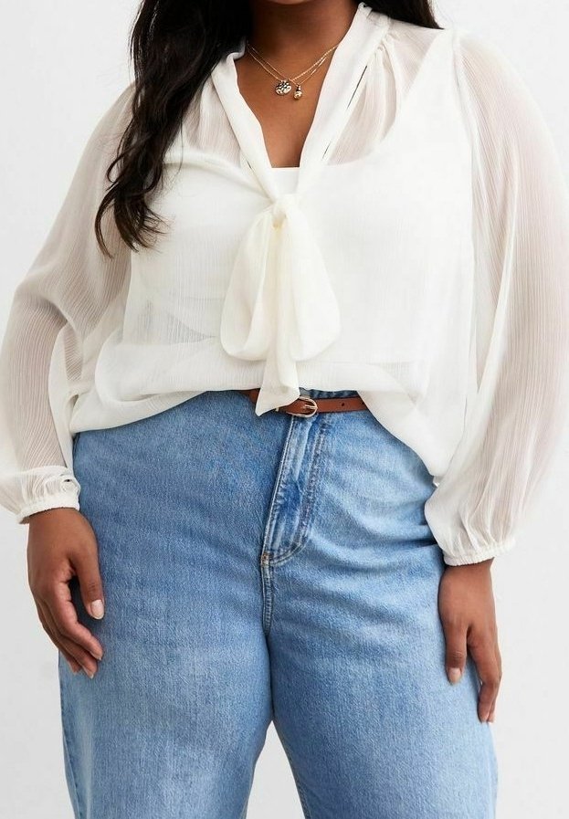 Femme portant un chemisier blanc transparent avec un nœud papillon au cou, rentré dans un jean taille haute bleu clair avec une ceinture marron.