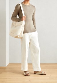 Beżowy sweter z dzianiny, off-white lniane spodnie, brązowe sandały na pasku i kremowa torba na ramię z kanwy z detalem guzika. Gładkie tekstury i neutralne kolory.