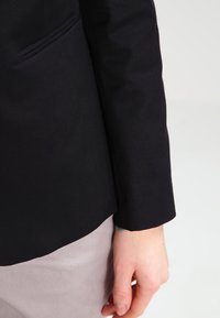 Ręka osoby spoczywa po boku, ubrana w czarną bluzę z długim rękawem i jasnoszare spodnie na jednolitym jasnym tle.