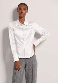 Vrouw in een witte satijnen blouse met knopen en grijze wijde broek, staand met één hand op haar heup tegen een effen achtergrond.