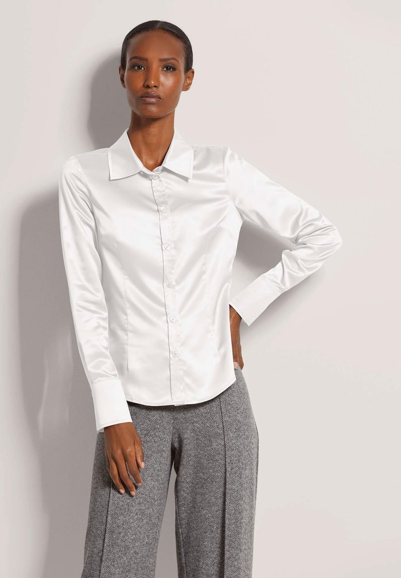 Vrouw in een witte satijnen blouse met knopen en grijze wijde broek, staand met één hand op haar heup tegen een effen achtergrond.