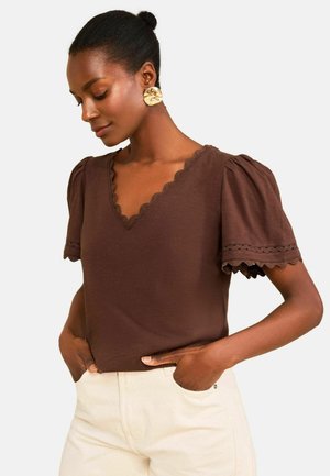 Sötét bőrű nő nagy arany fülbevalókkal, barna, hullámos V-nyakú felső puffos ujjakkal, világos színű nadrágban, kezét a zsebében tartva.