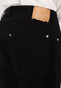 Pantaloni din denim negri cu cusături contrastante, accesorii din argint și o etichetă din piele maro pe talie cu detalii embossate.