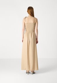 Lang beige jurk met een smocked lijfje, verstelbare schouderbanden en een vloeiende rok met een comfortabele pasvorm. Soepele textuur, minimale details.