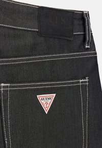 Jeans en denim noir avec des coutures blanches contrastantes, dotés d'un écusson triangulaire rouge et blanc "GUESS" sur la poche arrière.