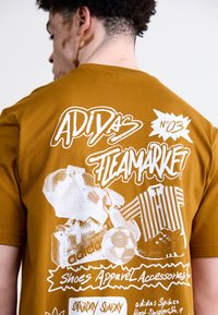 Barna pamut póló, fehér grafikával, amelyen az "ADIDAS FLEAMARKET" felirat és különböző stílusú cipők és kiegészítők képei láthatók.