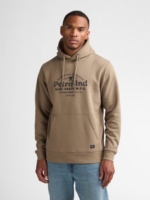 Giovane uomo che indossa una felpa con cappuccio color beige con testo e jeans azzurri chiari, in piedi contro uno sfondo chiaro e semplice.