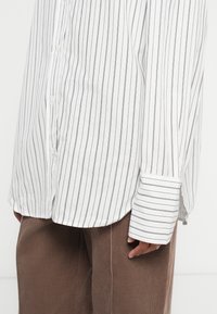 Witte lange mouwen overhemd met dunne zwarte verticale strepen, gedragen over een bruine corduroybroek, hand ontspannen naast het lichaam.