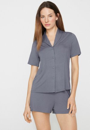 Frau mit langen braunen Haaren, die ein graues kurzärmeliges Hemd mit Knöpfen und passende Shorts vor einem schlichten Hintergrund trägt.