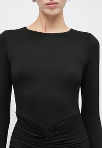 Γυναίκα που φοράει εφαρμοστή μαύρη μακρυμάνικη μπλούζα με στρογγυλή λαιμόκοψη και ρυτιδωτή λεπτομέρεια στη μέση, εμφανιζόμενη από το λαιμό μέχρι τους γοφούς.