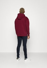 Felpa con cappuccio bordeaux a vestibilità comoda, in tessuto testurizzato con polsini a costine, abbinata a jeans skinny scuri e sneakers bianche.
