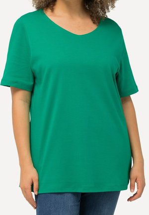 Kobieta ubrana w luźną zieloną koszulkę z krótkim rękawem i dekoltem w serek oraz niebieskie dżinsy na jednolitym jasnym tle.