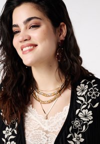 Gelaagde gouden en zilveren kettingen met een V-vormige hanger. Donkere kanten top met bloemrijke borduursels en grote oorbellen met rode edelstenen.