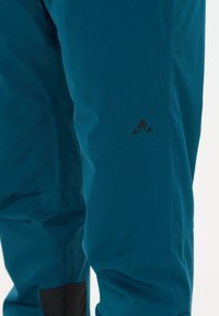 Nahaufnahme von tealfarbenen Outdoor-Hosen, die den Kniebereich mit kleinem schwarzem Berglogo und verstärktem schwarzem Stoff am Knöchel zeigen.