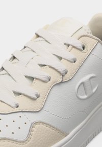 Κοντινό πλάνο λευκού δερμάτινου αθλητικού παπουτσιού με μπεζ ανάγλυφες λεπτομέρειες, λευκά κορδόνια, διάτρητη μύτη και ανάγλυφο λογότυπο Champion στο πλάι.