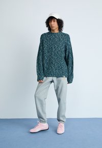 Suéter azul-esverdeado com manchas, de mangas compridas, combinado com calças de fato de treino cinza claro e sapatilhas desportivas rosa; modelo a usar um boné branco.