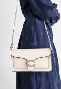 Beige leather crossbody bag with a smooth texture, silver chain strap, decorative crystal-accented clasp, and black stitching details.