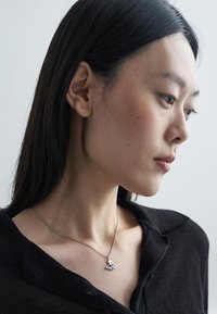 Ασημένιο κολιέ με τριγωνικό περικάρπιο που διαθέτει κοψίματα, φορεμένο σε λεπτή αλυσίδα. Το μοντέλο φοράει ένα μαύρο πουκάμισο με γιακά.